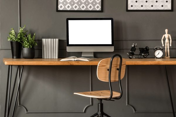 Dark open space living room interior with metal rack, grey armchair and plants in the background and study corner hairpin desk, books and empty monitor in the foreground